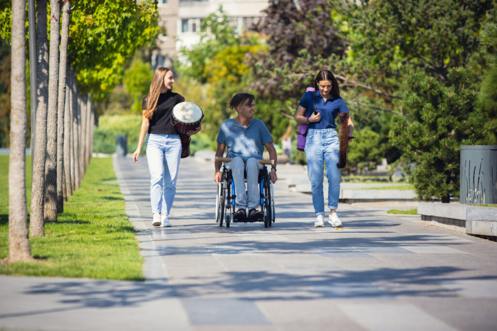 Three individuals enjoying an outdoor park path, one using a wheelchair, holding different musical instruments, highlighting inclusivity.